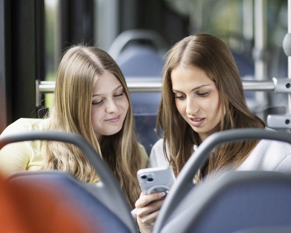 Zwei junge Frauen schauen im Bus zusammen auf ein Mobiltelefon 